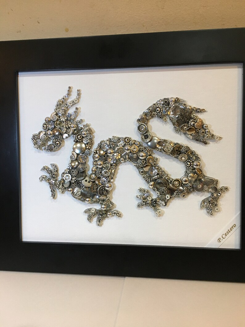 May include: A framed artwork depicting a silver dragon made of buttons and beads. The dragon is set against a white background and the frame is black. The artist's signature, "P. Cestero", is visible in the bottom right corner.