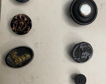 May include: A collection of vintage buttons in various shapes and sizes. The buttons are primarily black, with some featuring metallic gold and copper accents. Designs include floral patterns, geometric shapes, and a small oval button with a gold-colored reptile.