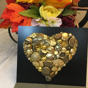 May include: A black framed heart-shaped artwork made of gold buttons. The buttons are arranged in a heart shape and are of various sizes and designs. The artwork is on a black background and has a wooden stand.