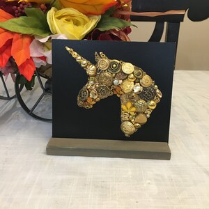 May include: A black square canvas features a unicorn head silhouette crafted from various gold and brass buttons. The artwork is mounted on a gray wooden stand, with a colorful floral arrangement in the background.