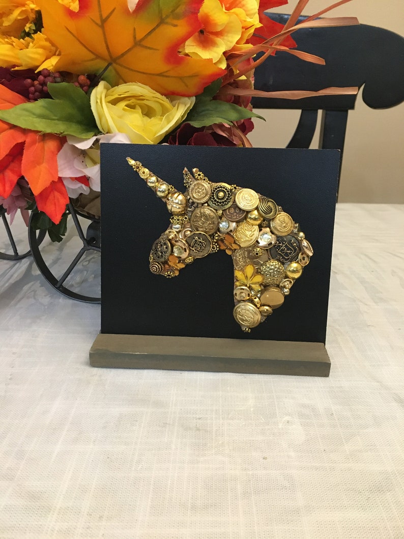 May include: A black square canvas features a unicorn head silhouette crafted from various gold and brass buttons. The artwork is mounted on a gray wooden stand, with a colorful floral arrangement in the background.