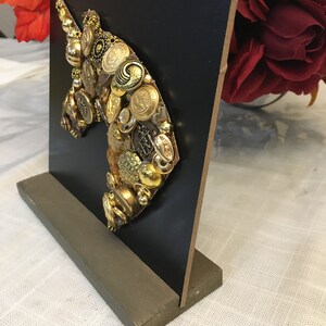 May include: A decorative unicorn head sculpture made from various gold-toned buttons and embellishments. The unicorn silhouette is mounted on a black square, supported by a gray rectangular base. The artwork is displayed on a white surface.