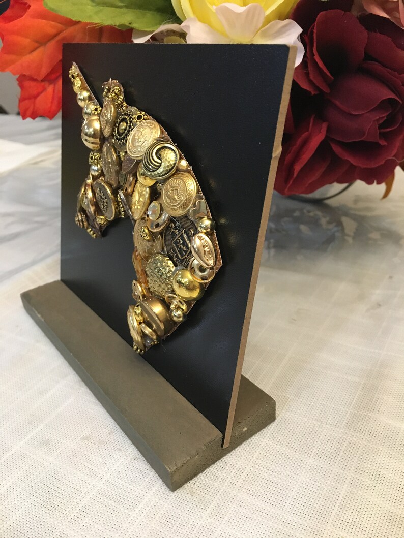 May include: A decorative unicorn head sculpture made from various gold-toned buttons and embellishments. The unicorn silhouette is mounted on a black square, supported by a gray rectangular base. The artwork is displayed on a white surface.