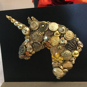 May include: A handcrafted artwork featuring a unicorn head silhouette. The design is composed of various gold-toned buttons and embellishments, set against a black background. The artwork is mounted on a wooden base.