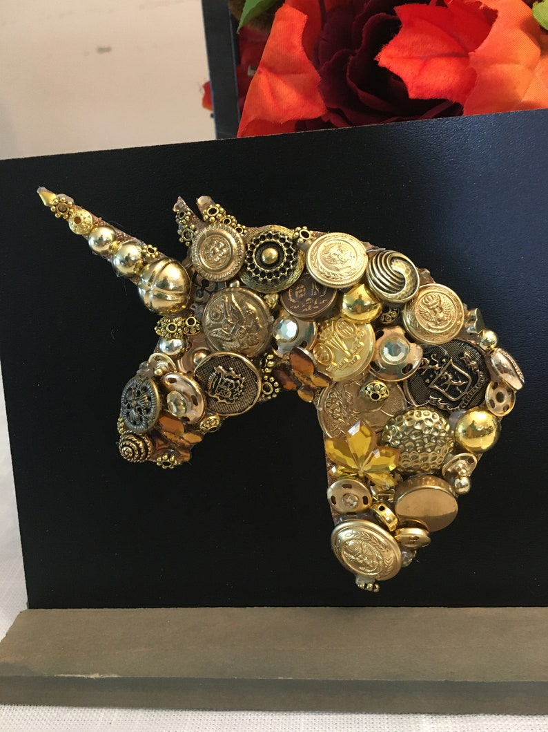May include: A handcrafted artwork featuring a unicorn head silhouette. The design is composed of various gold-toned buttons and embellishments, set against a black background. The artwork is mounted on a wooden base.