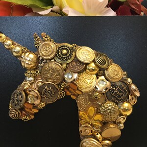 May include: A golden unicorn head made of various vintage buttons and embellishments. The buttons are in shades of gold, bronze, and brass, with some featuring intricate designs. The background is black, and the top has a floral arrangement.