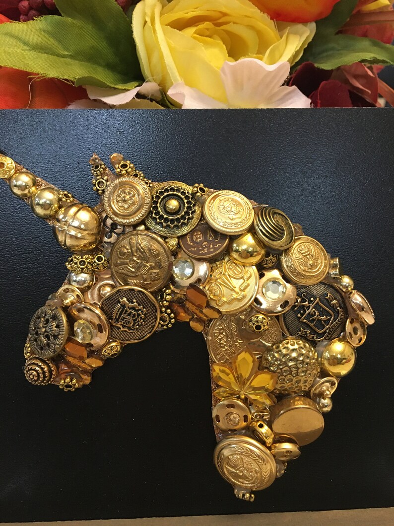 May include: A golden unicorn head made of various vintage buttons and embellishments. The buttons are in shades of gold, bronze, and brass, with some featuring intricate designs. The background is black, and the top has a floral arrangement.