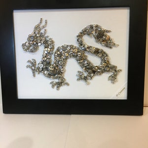 May include: A silver dragon made of buttons and other small metal objects is framed in a black frame. The dragon is on a white background. The signature "P. Castoro" is in the bottom right corner of the frame.