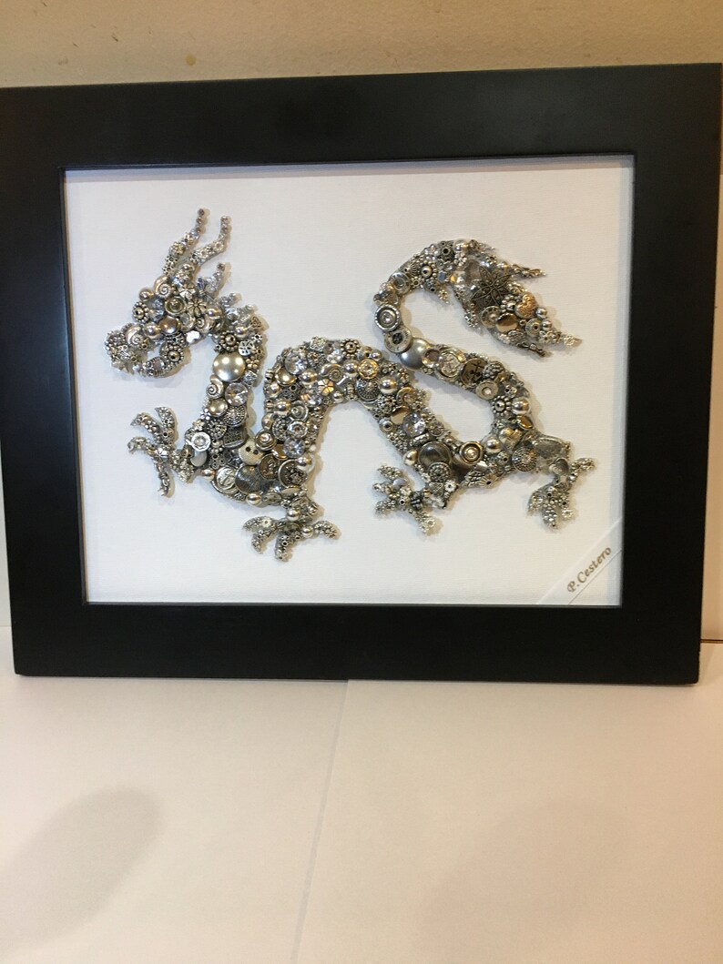 May include: A silver dragon made of buttons and other small metal objects is framed in a black frame. The dragon is on a white background. The signature "P. Castoro" is in the bottom right corner of the frame.