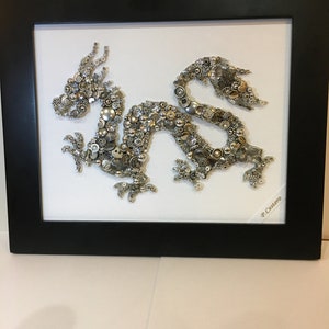 May include: A silver and gold dragon made of buttons and other small metal objects is framed in a black frame. The artwork is signed "P. Castero".