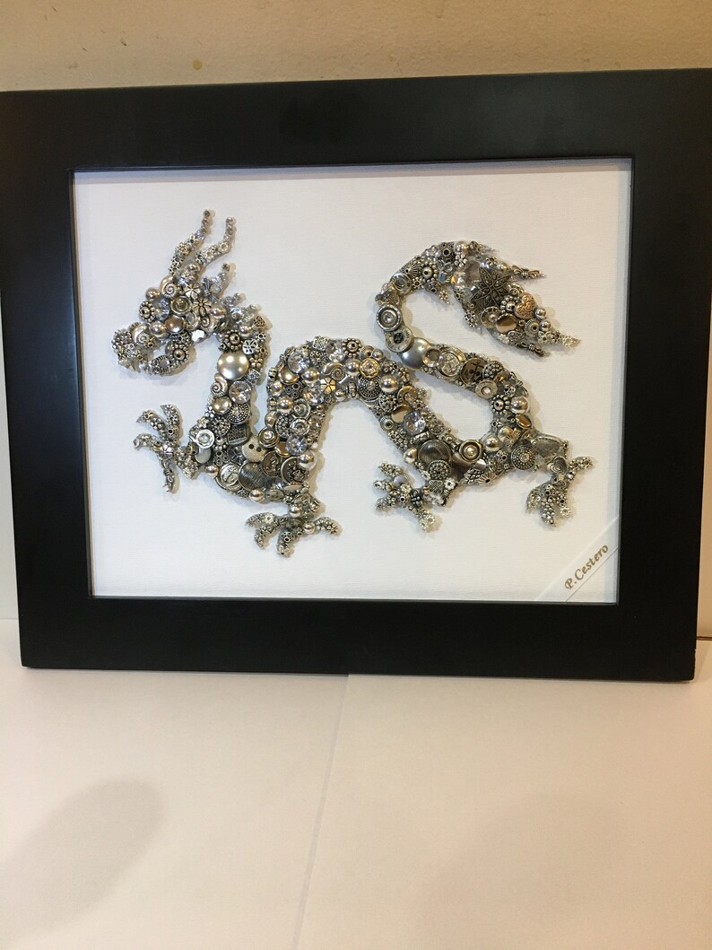 May include: A silver and gold dragon made of buttons and other small metal objects is framed in a black frame. The artwork is signed "P. Castero".
