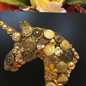 May include: A golden unicorn head crafted from various vintage buttons and embellishments, set against a black background. The buttons vary in size, shape, and texture, creating a dimensional effect. The artwork is a unique, handcrafted piece.