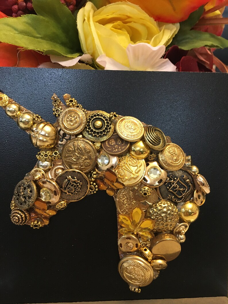 May include: A golden unicorn head crafted from various vintage buttons and embellishments, set against a black background. The buttons vary in size, shape, and texture, creating a dimensional effect. The artwork is a unique, handcrafted piece.