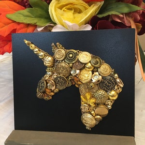 May include: A black square art piece featuring a unicorn head silhouette crafted from various gold-toned buttons. The buttons vary in size and design, creating a textured, dimensional effect. The artwork is mounted on a wooden base.