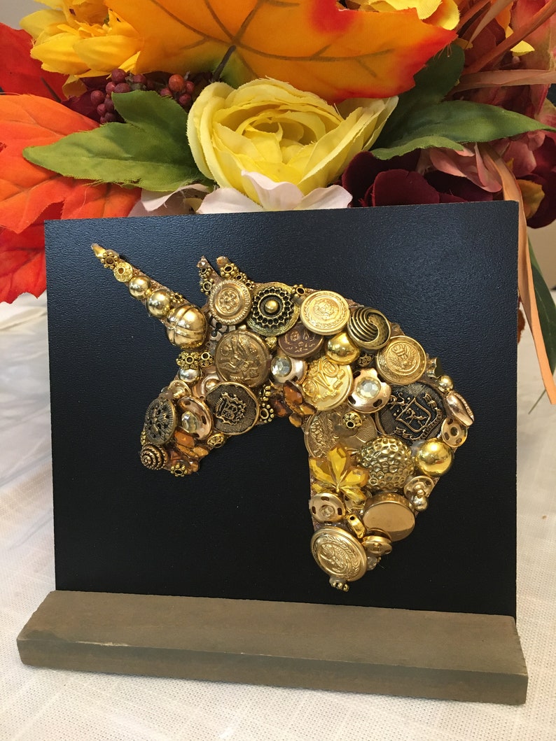 May include: A black square art piece featuring a unicorn head silhouette crafted from various gold-toned buttons. The buttons vary in size and design, creating a textured, dimensional effect. The artwork is mounted on a wooden base.