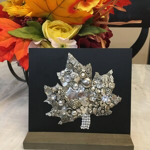 May include: A silver and black leaf-shaped art piece made of buttons, beads, and rhinestones. The piece is mounted on a black background and displayed on a wooden stand.