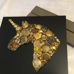 May include: A handcrafted artwork featuring a unicorn head silhouette. The unicorn is composed of various gold and brass buttons, beads, and embellishments, set against a black background. The piece is mounted on a square black base.