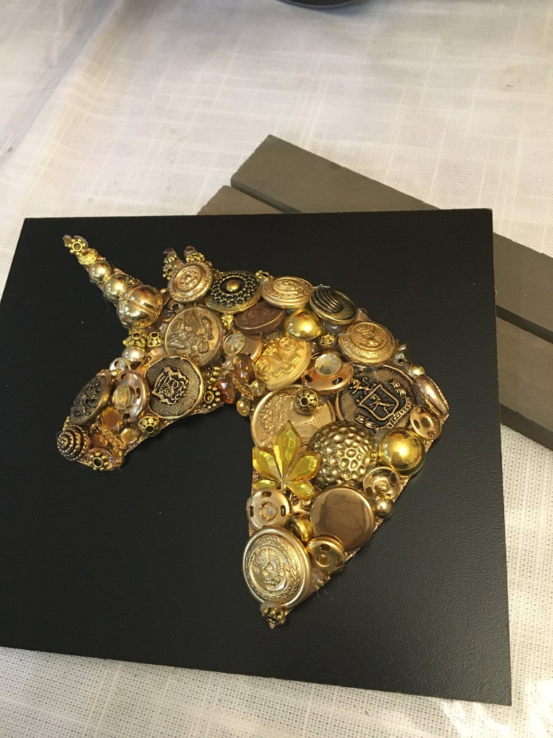 May include: A handcrafted artwork featuring a unicorn head silhouette. The unicorn is composed of various gold and brass buttons, beads, and embellishments, set against a black background. The piece is mounted on a square black base.
