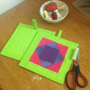 May include: Two green fabric pot holders with a pink and purple geometric design. One pot holder is folded in half. A pair of scissors and a spool of thread are on the table.