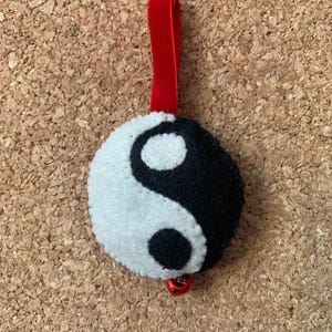 May include: A felt Yin and Yang ornament with a red velvet ribbon for hanging. The ornament is black and white with a small red bell attached to the bottom. The background is a corkboard.