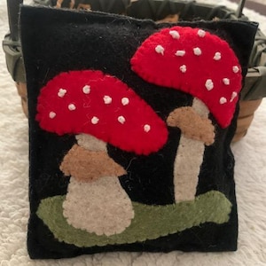 May include: A black felt pillow with two red and white mushrooms appliqued on it. The mushrooms have white spots and brown stems.