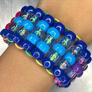 May include: A multi-strand beaded bracelet in shades of blue, clear, and purple. The bracelet is made of small, round beads strung together on elastic bands. The beads are a mix of solid colors and translucent with colorful designs inside.
