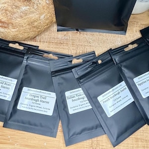 May include: Eight small black pouches with white labels containing sourdough starter cultures. The labels list the names of different sourdough starter types, including San Francisco, Alaskan Gold Rush, Oregon Trail, Italian Rustic, French Country, and  Appysian Sourdough.