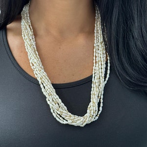 Puede incluir: Un collar de perlas de múltiples hilos con cuentas doradas. El collar está hecho de pequeñas perlas blancas y lo lleva una persona con el pelo largo y oscuro.