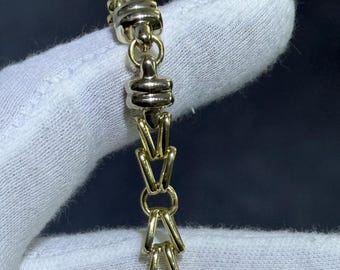 May include: A gold-toned bracelet with a unique link design. The bracelet features alternating sections of decorative, cylindrical links and V-shaped links. The clasp is a contrasting silver color. The bracelet is displayed against a dark background.
