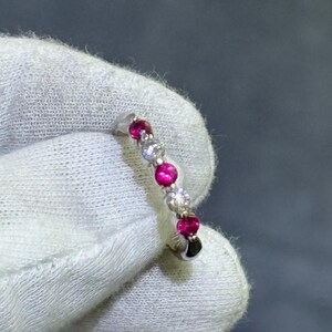 May include: A silver ring featuring a line of alternating red and clear gemstones. The ring is set against a white fabric, with a grey background.