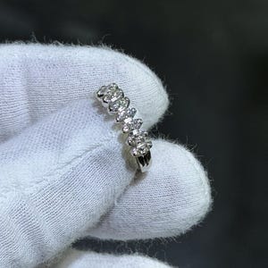 May include: A delicate silver ring featuring a series of small, sparkling gemstones. The ring's design showcases a row of evenly spaced, clear stones, creating a continuous band of brilliance. The ring is held against a white fabric background.