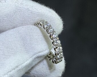 May include: A silver ring with a continuous row of round, clear gemstones. The ring is held against a white fabric background, showcasing the sparkle of the stones. The ring is a piece of jewelry.