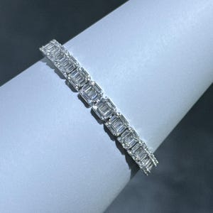 May include: A dazzling bracelet with a row of rectangular-cut gemstones. The gems are set closely together, forming a continuous line of sparkle. The bracelet is shown on a light grey surface, accentuating its elegant design.