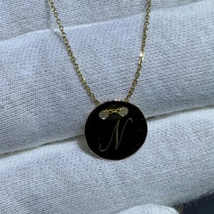 Puede incluir: Un collar dorado con una cadena fina y un colgante circular. El colgante negro muestra la letra "N" en cursiva. El collar se presenta sobre una superficie blanca.