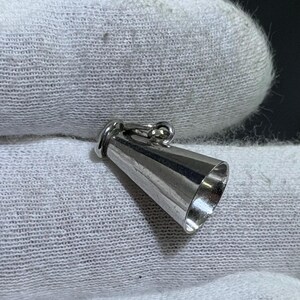 May include: A small, polished silver-toned metal charm shaped like a megaphone or cone. The object features a wide, open circular end and a narrow closed end with a small attached jump ring for suspension, displaying a smooth, reflective surface.