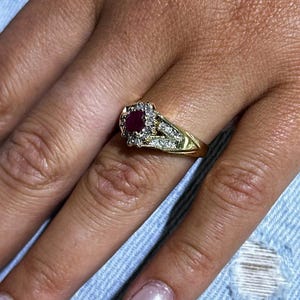 以下が含まれることがあります： 赤いルビーの宝石を小さなダイヤモンドで囲んだ金の指輪。指輪は繊細なデザインで、シャンクが分かれています。