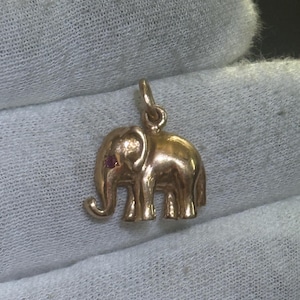May include: A gold-colored elephant charm with a small red gem eye. The charm is detailed with visible tusks, ears, and legs. The elephant is a popular symbol of good luck and fortune. This charm is suitable for a necklace or bracelet.