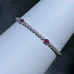 May include: A delicate silver bracelet featuring alternating round and oval-cut gemstones. The bracelet has three oval-cut, vibrant red gemstones and numerous smaller, clear round gemstones. The bracelet is displayed on a white surface.
