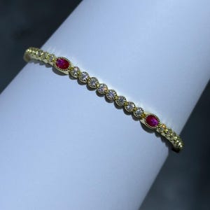 May include: A gold-colored bracelet with alternating oval red gemstones and round clear gemstones. The bracelet is set on a white surface and has a delicate, ornate design.