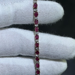 Może przedstawiać: Złota bransoletka z naprzemiennymi owalnymi kamieniami szlachetnymi z rubinu i małymi, okrągłymi, przezroczystymi kamieniami szlachetnymi. Bransoletka jest prezentowana na białym tle.