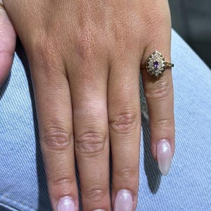 Könnte beinhalten: Ein goldener Ring mit einem violetten Edelstein, der von kleineren, klaren Edelsteinen umgeben ist. Der Ring wird an einem Finger mit hellrosa Nagellack getragen.