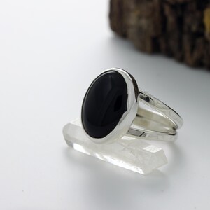 May include: A silver ring with a large, oval black stone. The ring has a hammered texture on the band and is displayed on a clear crystal. The ring is a piece of jewelry.