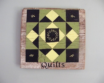 May include: A wooden sign with a black, yellow, and green quilt block pattern painted on it. The sign says "Quilts" at the bottom.