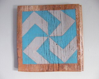 May include: A wooden square sign with a geometric pattern painted in teal and gray. The sign has a rustic, distressed look and is framed with a brown border.