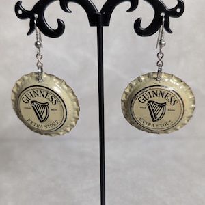 May include: Earrings made from Guinness Extra Stout bottle caps. The earrings are round, with the Guinness logo and text in black and white. They hang from silver-colored ear wires and are displayed on a black metal earring stand.
