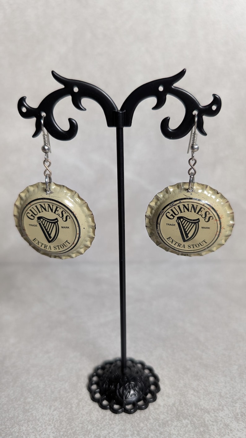 May include: Earrings made from Guinness Extra Stout bottle caps. The earrings are round, with the Guinness logo and text in black and white. They hang from silver-colored ear wires and are displayed on a black metal earring stand.