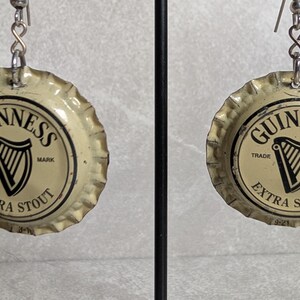 May include: A pair of earrings made from Guinness Extra Stout bottle caps. Each cap features the Guinness logo with a harp and the words "Trade Mark" and "Extra Stout" in black on a cream background. The earrings hang from silver-colored hooks.