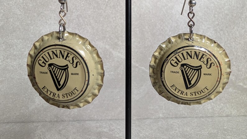 May include: A pair of earrings made from Guinness Extra Stout bottle caps. Each cap features the Guinness logo with a harp and the words "Trade Mark" and "Extra Stout" in black on a cream background. The earrings hang from silver-colored hooks.