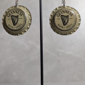 May include: A pair of earrings made from Guinness Extra Stout bottle caps. The earrings are round with a silver-colored edge and feature the Guinness logo with a harp and the words "Extra Stout". They hang from silver-colored hooks.