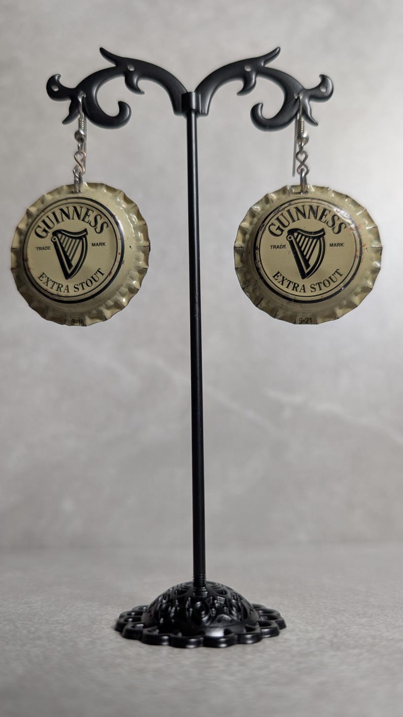May include: A pair of earrings made from Guinness Extra Stout bottle caps. The earrings are round with a silver-colored edge and feature the Guinness logo with a harp and the words "Extra Stout". They hang from silver-colored hooks.